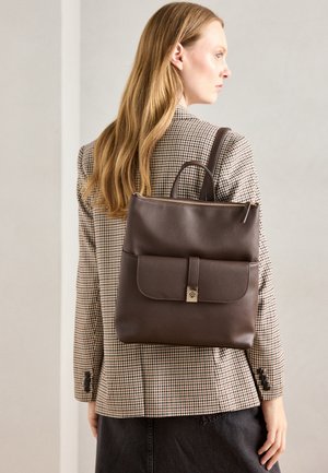 Smeđa kožna ruksak s preklopnim džepom, glavnim zip zatvaračem i dvostrukim ručkama, nošen preko kariranog sakoa s tamnom suknjom.