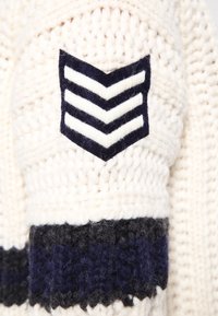 Neulepaita kreemissä, jossa on laivastonsininen chevron-patch ja tummansininen nauha. Teksturoitu paksulla langalla; pehmeä villamateriaali.