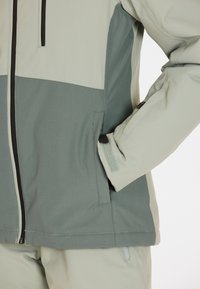 Leichte, zweifarbige Jacke in Hellgrün und Dunkelgrün, mit strukturiertem Stoff, Reißverschlusstaschen und verstellbaren Bündchen mit Klettverschluss.