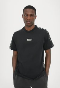 Μαύρη μπλούζα με κοντά μανίκια, με υ�φασμάτινες λεπτομέρειες στους ώμους, λαιμόκοψη τύπου crew και ένα εκτυπωμένο λογότυπο μπροστά.