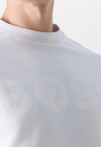 Gros plan sur une personne portant un sweat-shirt blanc avec des lettres en relief formant le mot "BOSS" sur la poitrine.