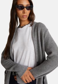 Cárdigan gris de punto con abertura frontal y patrón texturizado, llevado sobre una camiseta blanca. El modelo lleva accesorios como gafas de sol oscuras y un collar de cadena.