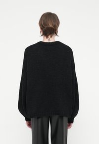 Maglione nero lavorato a maglia con una vestibilità rilassata, spalle basse e scollatura a costine. Tessuto texturizzato con un intreccio morbido, abbinato a pantaloni neri lisci.