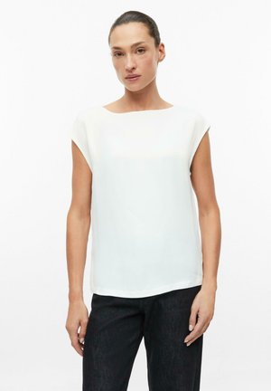 Donna con capelli scuri legati indietro che indossa una canotta bianca senza maniche e pantaloni neri su uno sfondo bianco uniforme.