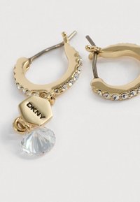 Zlaté kruhové náušnice so svetlými kryštálmi a visiaceho krištáľového prívesku. Sú vybavené logom DKNY na šestuholníkovej značke. Hladká textúra.