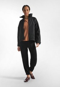 Musta kevyt takki korkealla kauluksella, päällä ruskea paita ja mustat housut. Asun viimeistelevät tummanpunaiset nyörikengät.