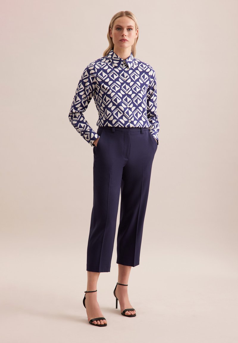 Camicia a maniche lunghe di colore blu navy con un motivo geometrico bianco, abbinata a pantaloni tailored blu navy e tacchi neri a strappo. Tessuto dalla texture liscia.