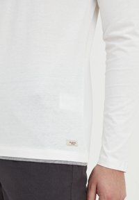 Hvid langærmet t-shirt lavet af blødt stof, med ribbet krave, buet bund og et lille "BLEND" label på siden.