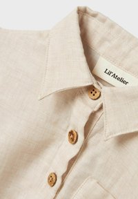 Camicia di lino beige chiaro con colletto a punta, bottoni in sughero e un'etichetta con la scritta "Lil'Atelier". La texture è tessuta e morbida.