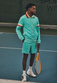 Meeste tennisetreening: türkiissinine tõmbluku peas jakiga, punaste ja kollaste aktsentidega, sobivad lühikesed püksid, valged tossud mustade detailidega, hoides oranži reketit.