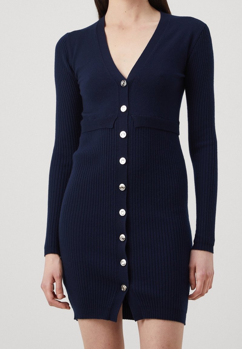Femme portant une robe en tricot côtelé bleu marine ajustée avec manches longues et boutons argentés sur le devant, sur un fond uni.