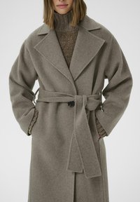 Abrigo de mezcla de lana gris con cinturón de lazo y grandes solapas. El diseño cuenta con mangas anchas y un suéter marrón texturizado debajo.