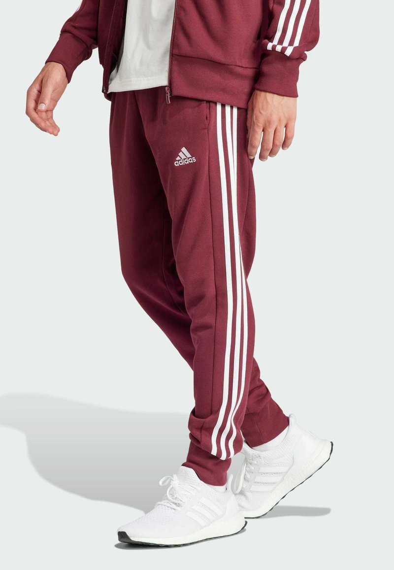 Burgundijas sporta bikses ar baltām trim svītrām un izšūtu logo uz kreisā augšstilba, izgatavotas no gludas auduma.