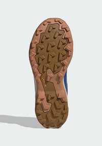 Sole běžecké obuvi na trail ve hnědé a růžové gumě s vícesměrnými špunty pro trakci. Obsahuje logo značky v oranžové barvě a texturovaný grip.