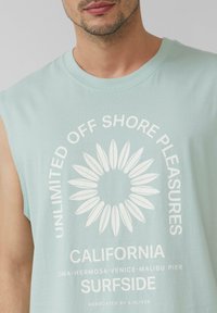Mintgrünes ärmelloses T-Shirt mit einem weißen kreisförmigen Grafikelement, das Surfbrettformen und Text zeigt, aus weichem Baumwollstoff.