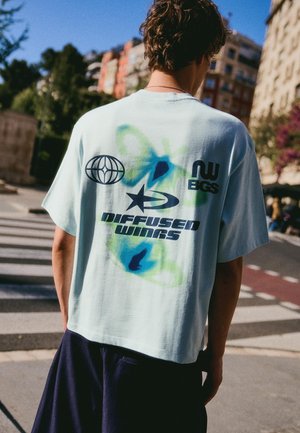 Giovane con capelli mossi che indossa una maglietta azzurro chiaro oversize con farfalla e loghi, in piedi sulle strisce pedonali di una strada di città alla luce del giorno.