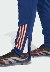 Die marineblauen Sporthosen verfügen über drei vertikale orangefarbene Streifen an der Seite und einen Reißverschluss am Saum, kombiniert mit schwarzen und roten Schuhen.