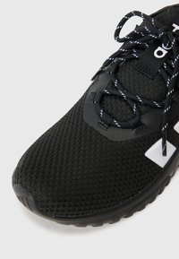 Chaussure de sport noire avec une tige en maille texturée, dotée de lacets noirs et blancs et d'une semelle en caoutchouc. Branding et accents notables dans des couleurs contrastées.