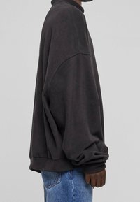 Persona con una sudadera negra de manga larga y suelta y jeans de mezclilla azules, vista de perfil sobre un fondo gris liso.