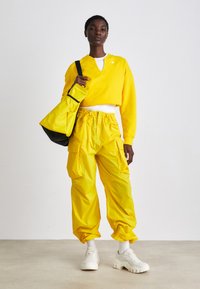 Sárga, cropped pulóver, hozzá illő cargo nadrág és fehér sneakers. Tote táska fekete kiegészítőkkel. Simább felületek, laza fazon és állítható derékpánt.