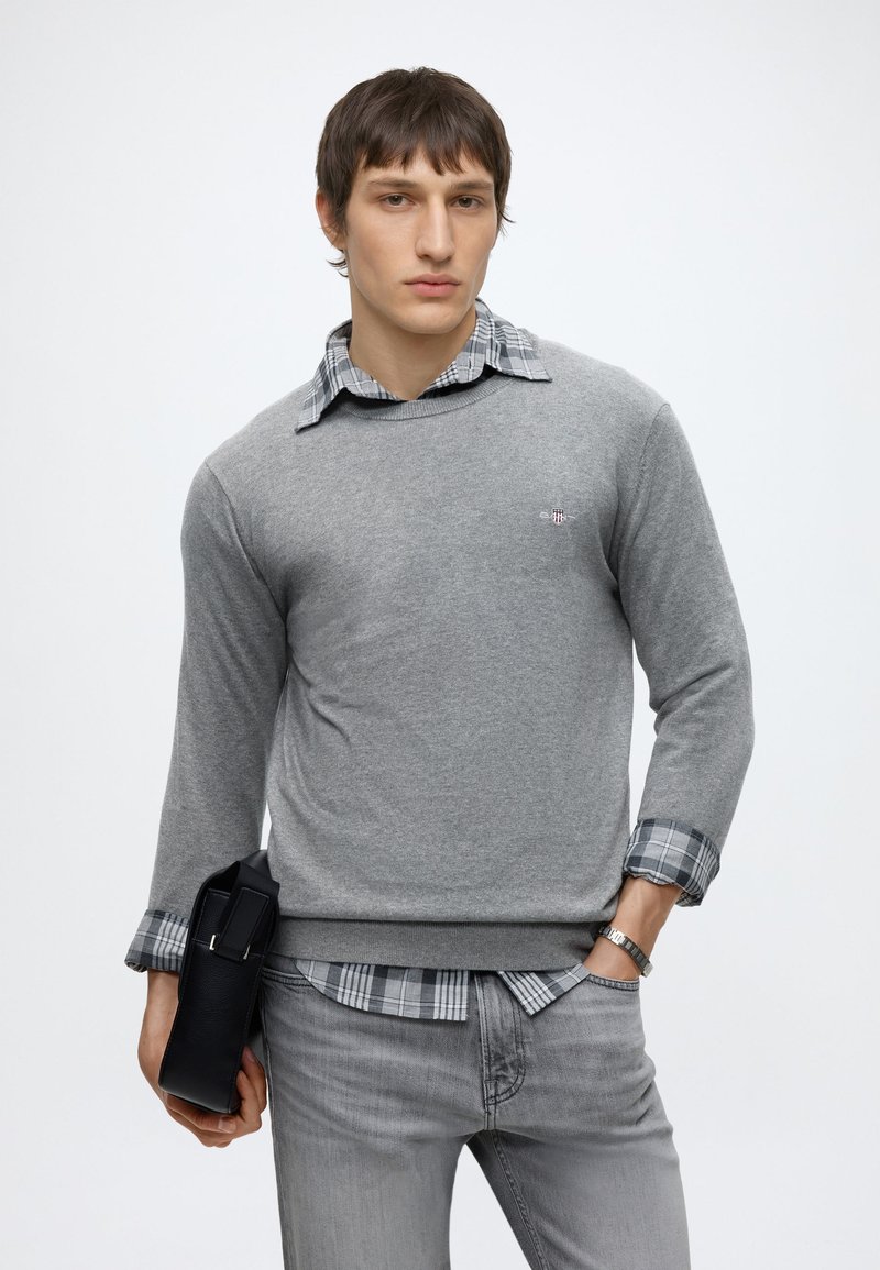 Young man wearing a gray sweater over a plaid shirt and gray jeans, holding a black bag in his left hand, posing against a white background.