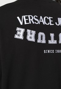 Černá mikina s bílým vyšitým nápisem „VERSACE JEWELTURE“ a „SINCE 1980“. Látka má hladkou strukturu.