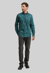 Teal gemusterde blouse met een button-down kraag en een borstzak, gecombineerd met donkergrijze jeans en zwarte laarzen, staand tegen een neutrale achtergrond.