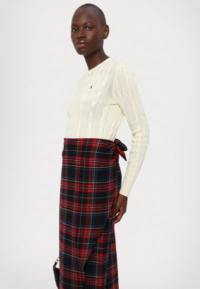Maglione crema a trecce abbinato a una gonna a pieghe in plaid nera e rossa. L'outfit presenta un accento legato in vita.