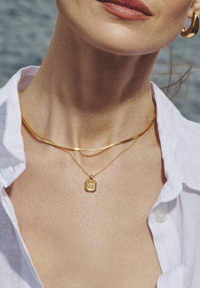 Joias de ouro incluem uma corrente plana e elegante e um pendente delicado com uma forma retangular que apresenta um design de explosão solar. Camisa branca ao fundo.