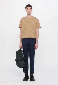 Brown cotton t-shirt with embossed logo, navy slim-fit pants, black leather shoes, and a dark gray backpack with adjustable straps.