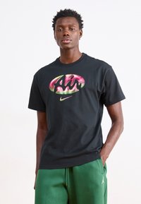 Svart bomull T-shirt med en färgglad "Air"-logotyp och Nike swoosh. Modell bär gröna träningsbyxor. Enkel design med rund hals.