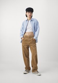Chemise boutonnée rayée bleu clair, crop top blanc, pantalon en velours côtelé marron à jambes larges et baskets grises avec des semelles blanches. Casquette noire et accessoires.