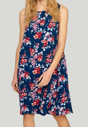 Femme portant une robe sans manches bleu marine avec un motif floral rouge, rose et blanc sur un fond uni clair.