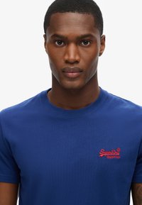 Camiseta de algodón azul royal con cuello redondo y mangas cortas. Presenta un pequeño logo bordado rojo en el lado izquierdo del pecho.