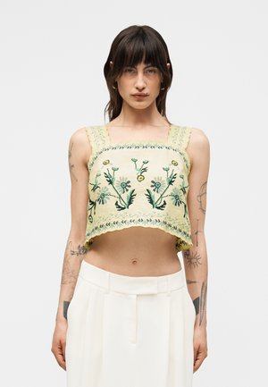 Kvinde med mørkt hår iført en gul kort top med blomstret broderi og hvide højtaljede bukser, stående mod en ensfarvet baggrund.