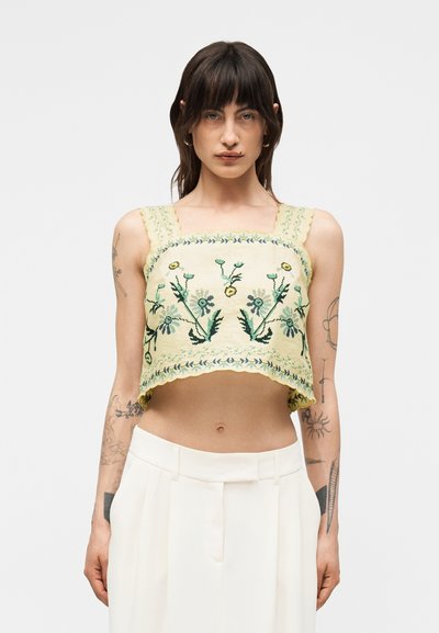 Woman with dark hair wearing a yellow cropped top with floral embroidery and white high-waisted pants, standing against a plain background.