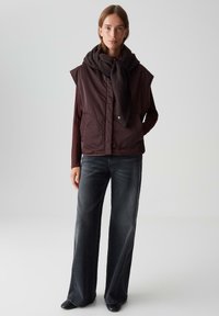 Braune gepolsterte Weste mit hohem Kragen, großen seitlichen Taschen, über einem weinroten Langarmoberteil getragen, kombiniert mit weit geschnittenen dunklen Jeans.