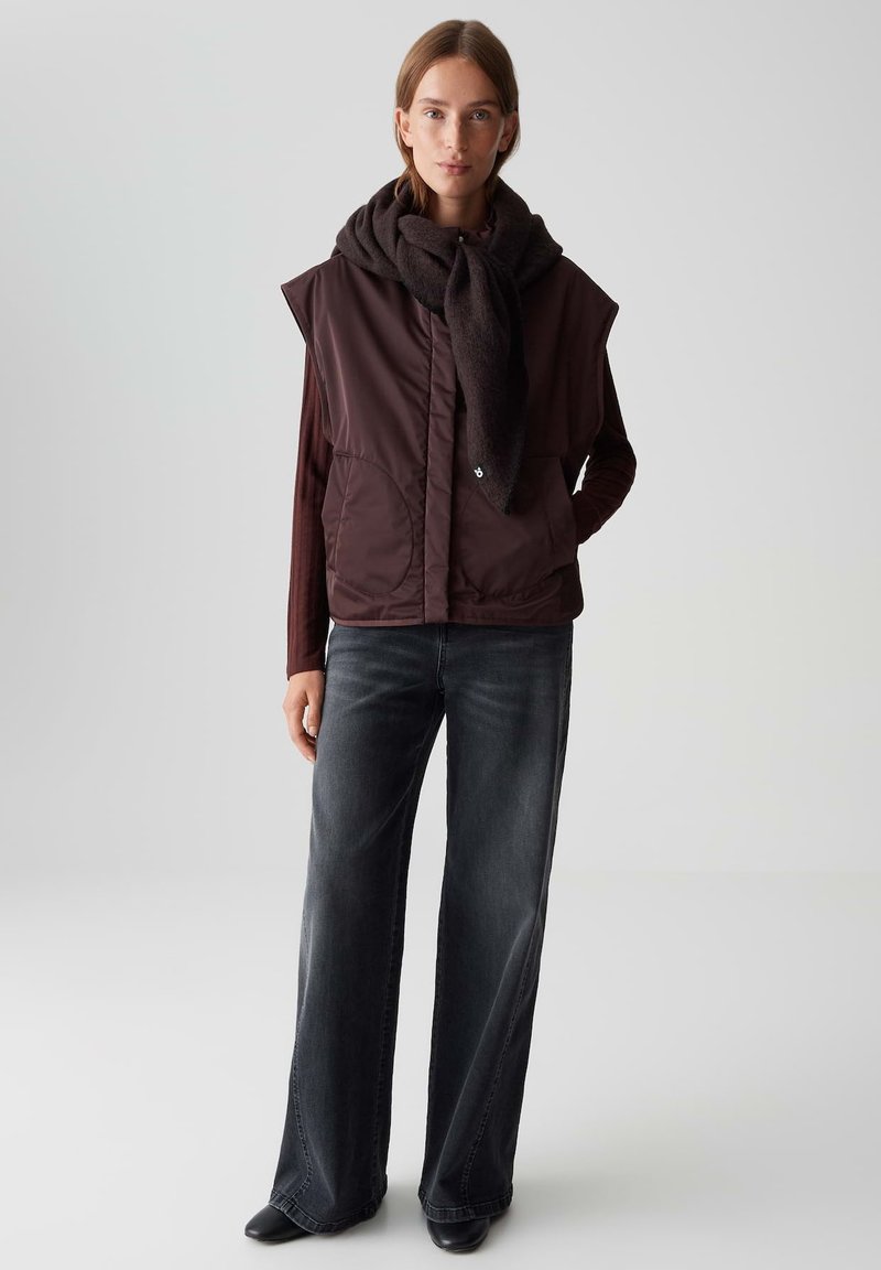 Braune gepolsterte Weste mit hohem Kragen, großen seitlichen Taschen, über einem weinroten Langarmoberteil getragen, kombiniert mit weit geschnittenen dunklen Jeans.