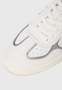 Vit sneakerspets med krämfärgade snören, perforerade läderpaneler och metallicsilverdetaljer på en ljusfärgad sula.