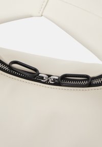 Bolso de cuero color crema con forma curva, que presenta una cremallera negra y herrajes plateados. Superficie texturizada con costuras visibles.