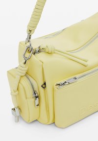 Żółta skórzana torebka z trzema zamykanymi na zamek przegrodami oraz uchwytem akcentowanym liną z srebrnymi zapięciami, na białym tle.