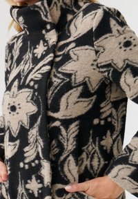 Chaqueta de forro polar en negro y beige, con patrones florales, un cuello alto y bolsillos laterales. Textura suave con un diseño de corte relajado.