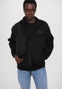 Giacca nera con zip e dettagli a coste, abbinata a una borsa a vita nera slim. Il modello indossa una maglietta nera e jeans azzurri chiari.