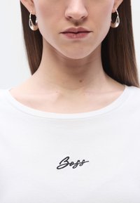 Λευκό βαμβακερό t-shirt με στρογγυλή λαιμόκοψη και μαύρο κεντητό κείμενο "Boss" στο στήθος, συνδυασμένο με ασημένι�α σκουλαρίκια κρίκους.