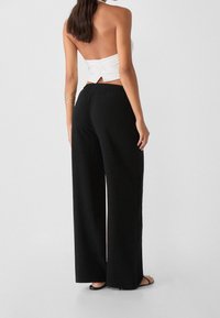 Weißes Neckholder-Top mit Rückenschnürung, kombiniert mit weit geschnittenen schwarzen Hosen. Glatte Stoffstruktur und schlichtes Design, dazu flache Sandalen.