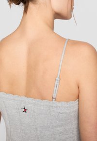 Grå strikket topp med bølget utskjæring, tynne justerbare stropper og dekorativ stjernedetalj med røde og hvite detaljer. Teksturerte mønstre er synlige.