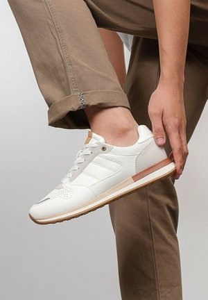 Personne portant un pantalon beige, pliant la jambe pour tenir une chaussure blanche avec des accents beige et marron sur la semelle.