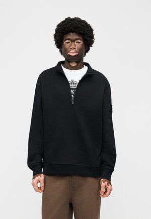 Mlada oseba z vitiligom stoji, oblečena v črno pulover z zadrgo do polovice in rjave hlače pred enobarvnim ozadjem.