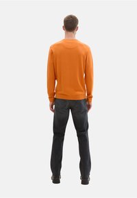 Suéter naranja de manga larga, con puños y cuello acanalados, combinado con jeans oscuros. La vista trasera muestra una silueta ajustada y un diseño minimalista.