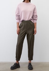 Lichtpaarse trui met geribbelde mouwen, gecombineerd met olijfgroene gestreepte joggingsbroek met elastische tailleband en zijzakken. Zwarte schoenen.